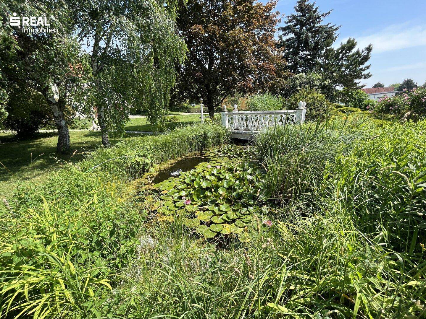 Malerischer Gartenteich mit Seerosen und einer dekorativen Brücke, umgeben von üppigem Grün.