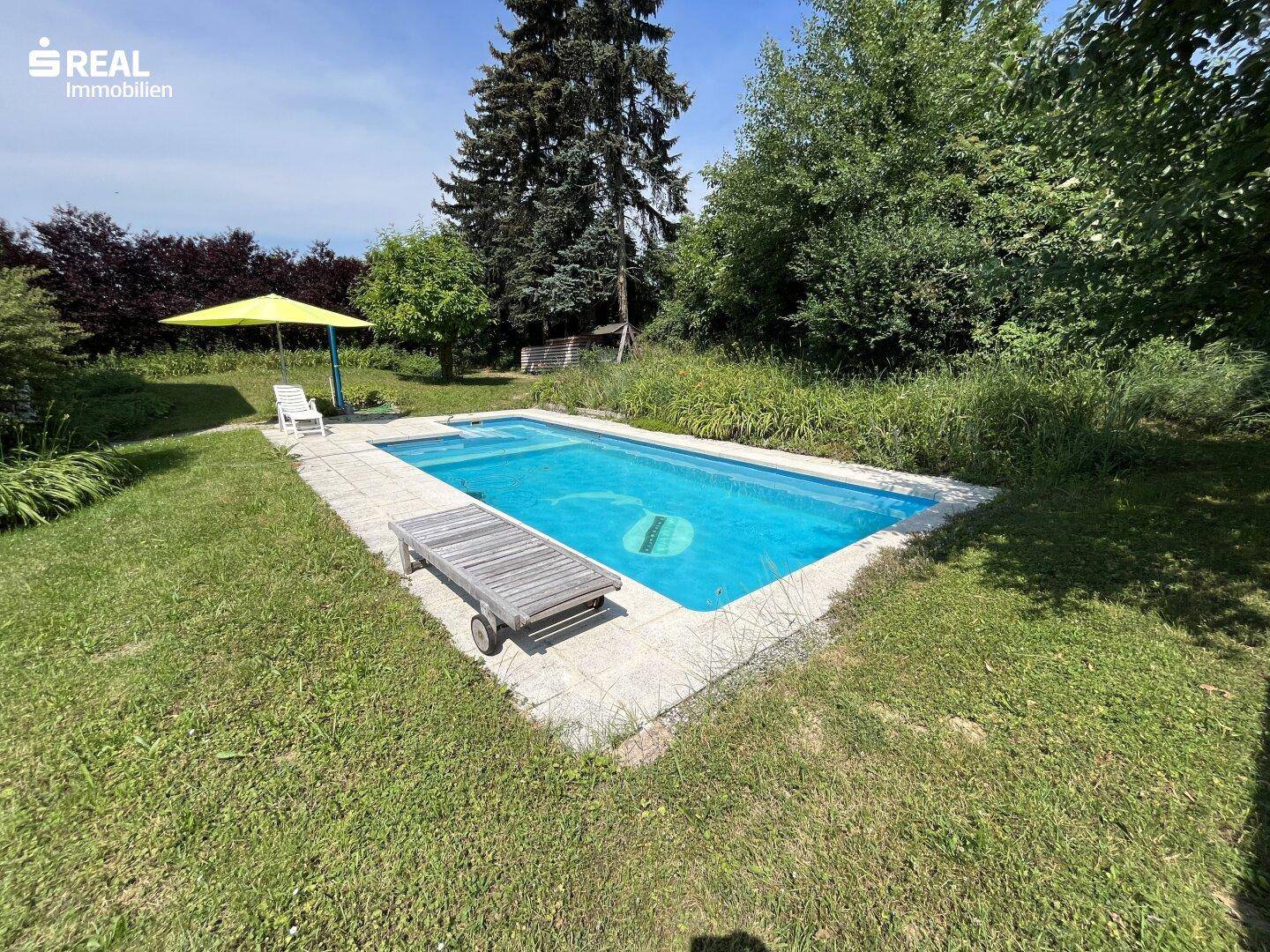 Rechteckiger Swimmingpool im Gartenbereich mit Liegefläche und Sonnenschirm unter blauem Himmel.