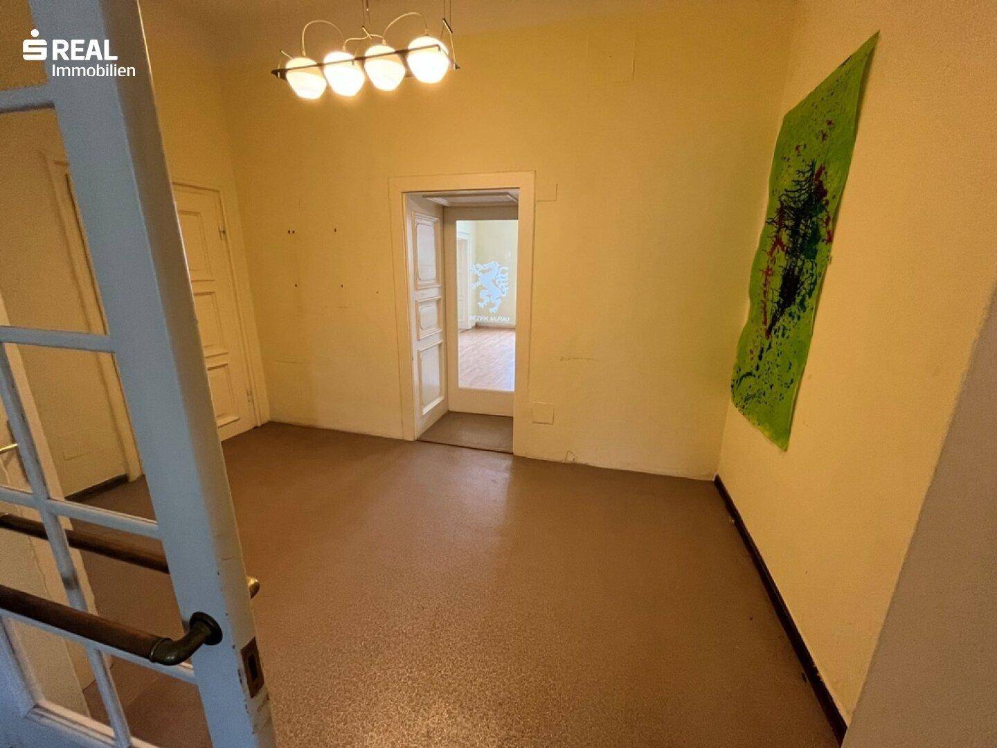 Leerer Raum mit einfacher Beleuchtung und Zugang zu weiteren Zimmern durch eine Glastür.