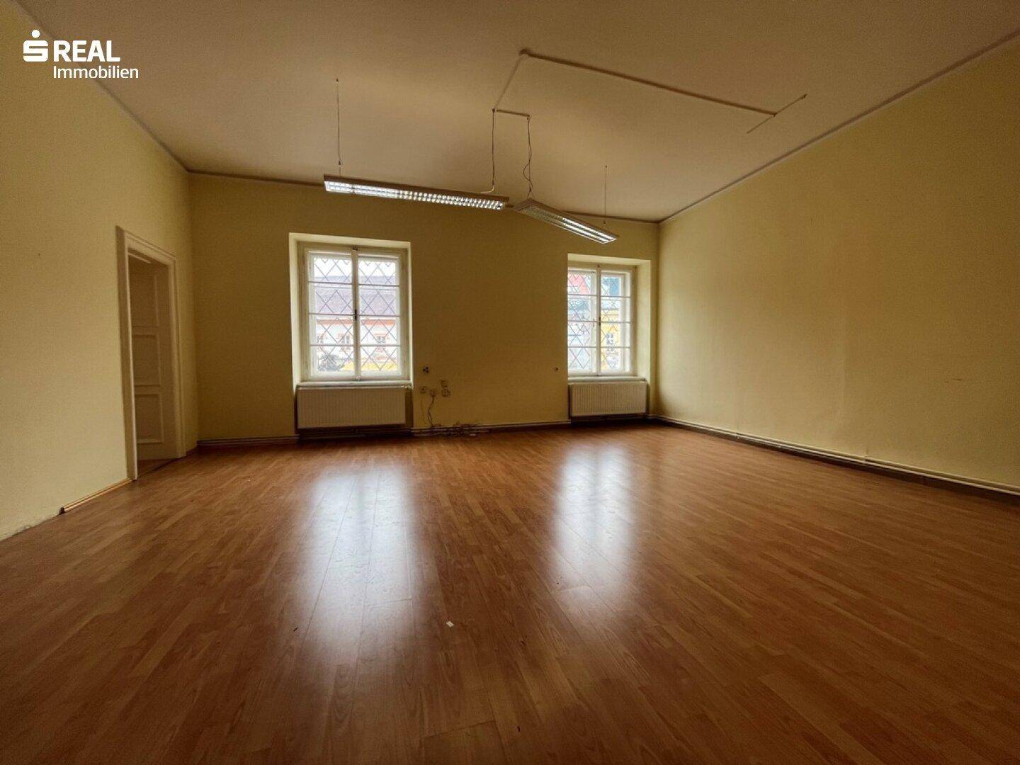 Geräumiger, leerer Raum mit Laminatboden und zwei großen Fenstern, die viel Tageslicht hereinlassen.