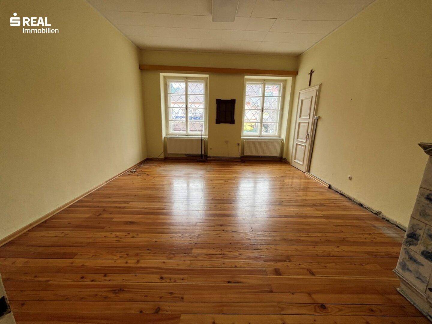 Großer Raum mit Holzdielenboden und zwei Fenstern, die den Blick auf die Umgebung freigeben.