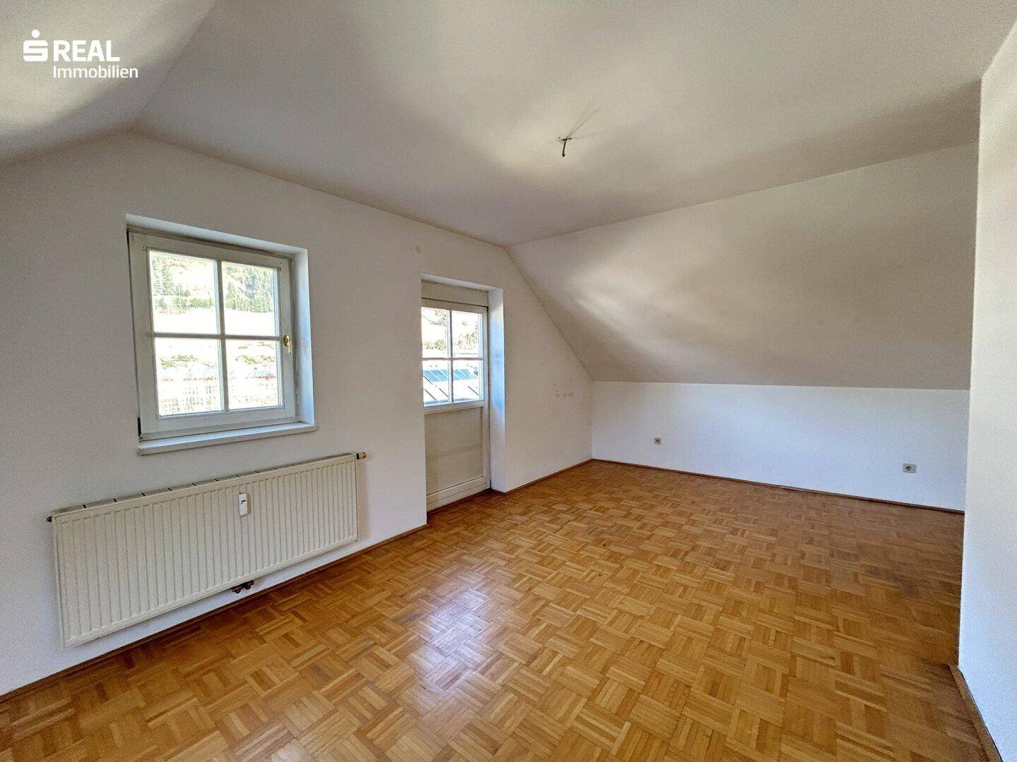 Helles Wohnzimmer mit Parkettboden, Heizkörper und zwei Fenstern.