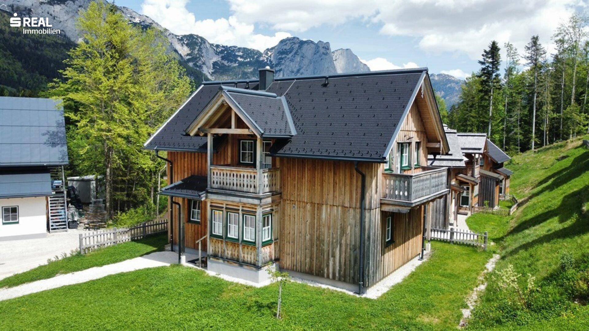 Alpines Holzhaus mit dunklem Dach und Balkonen, umgeben von grünen Wiesen und majestätischen Bergen.