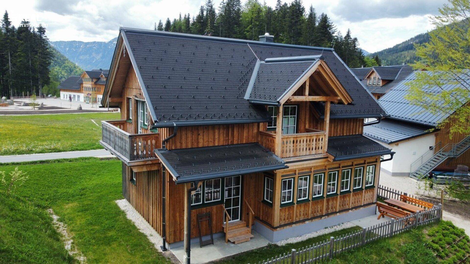 Traditionelles Holzhaus mit dunklem Dach und Balkonen, umgeben von grünen Wiesen und bewaldeten Bergen.
