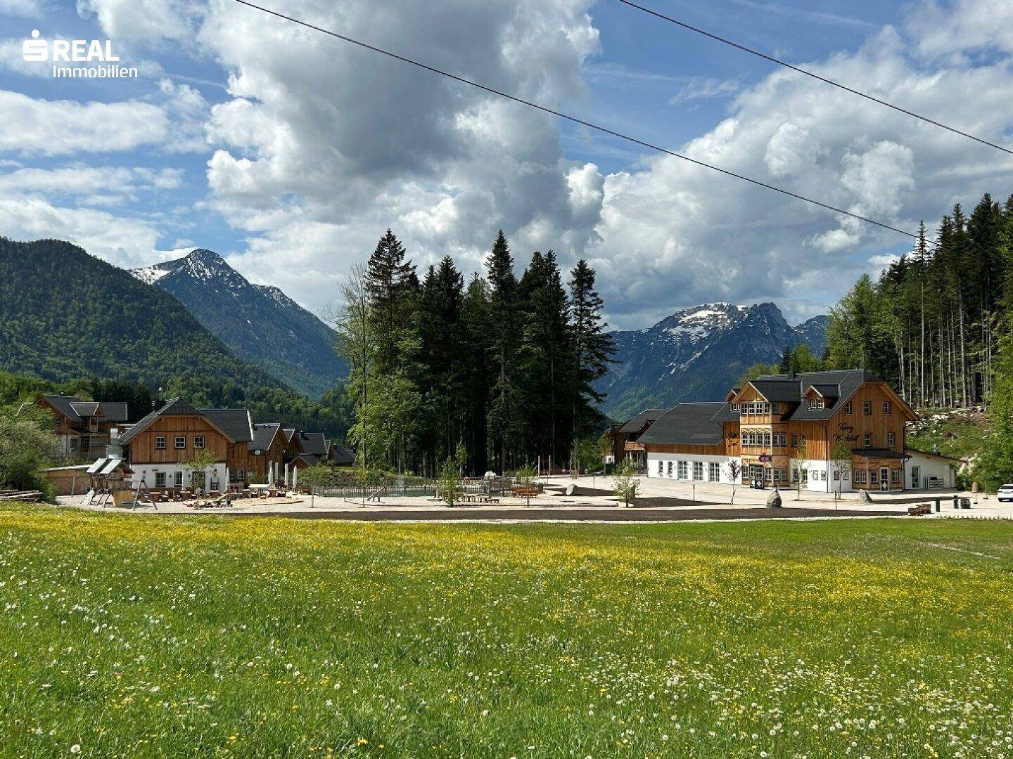 Weitläufige Landschaft mit blühenden Wiesen, traditionellen Gebäuden und beeindruckenden Bergen im Hintergrund.