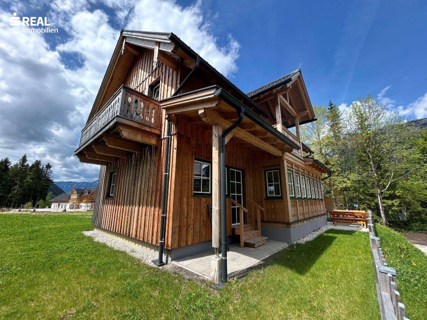 Seitenansicht eines Holzhauses mit überdachtem Eingangsbereich und Balkonen, umgeben von Natur.