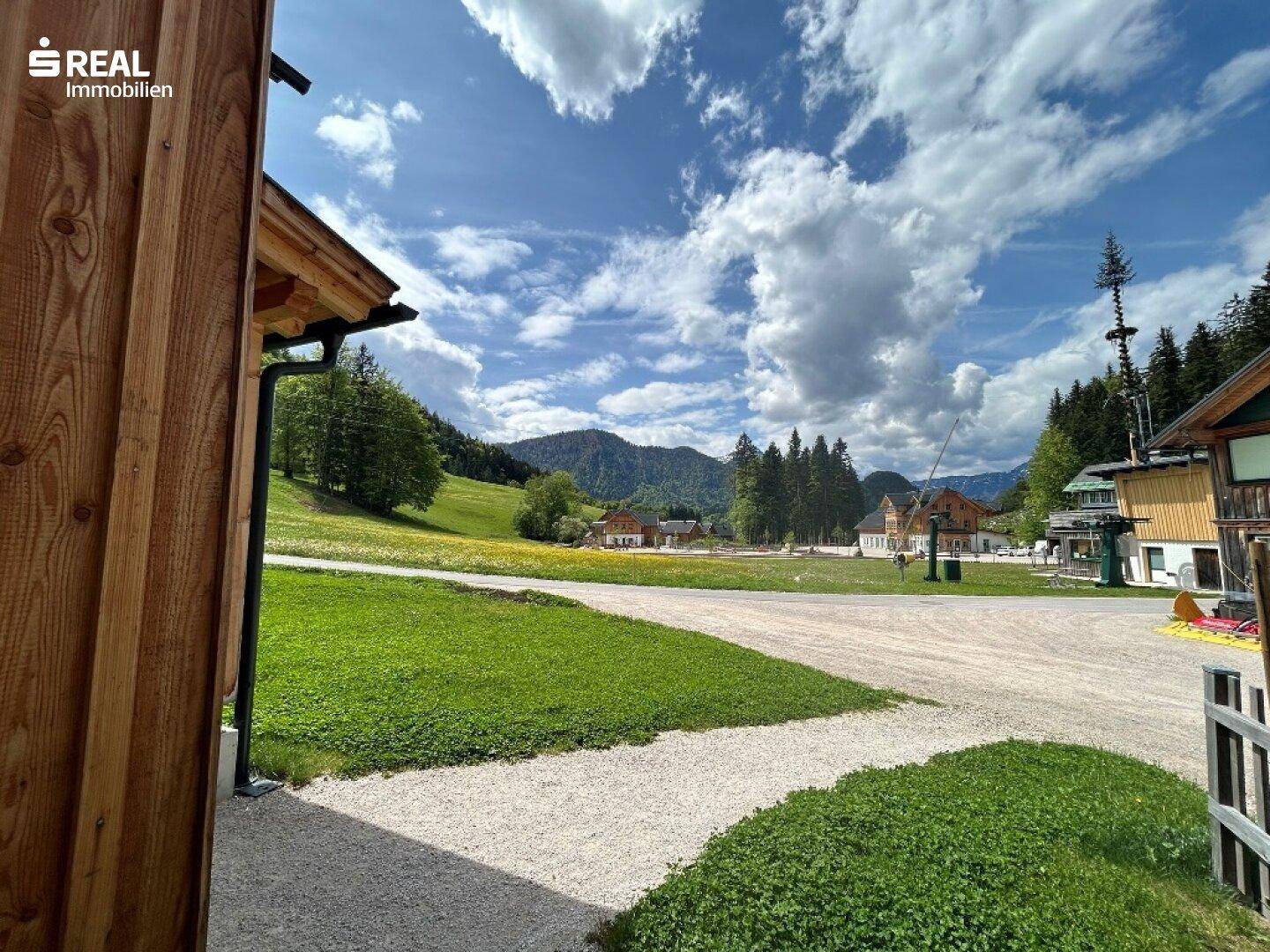 Blick von der Unterkunft auf eine weite Wiese und die umliegende Berglandschaft unter blauem Himmel.