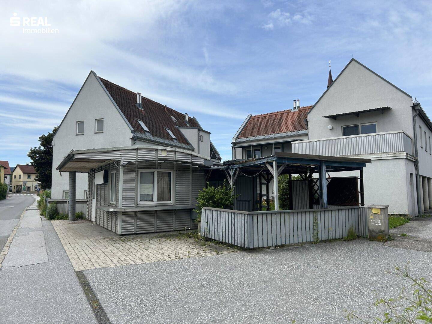 Ansicht von zwei Gebäuden mit einem kleinen Anbau und einem überdachten Bereich an einer Straße.