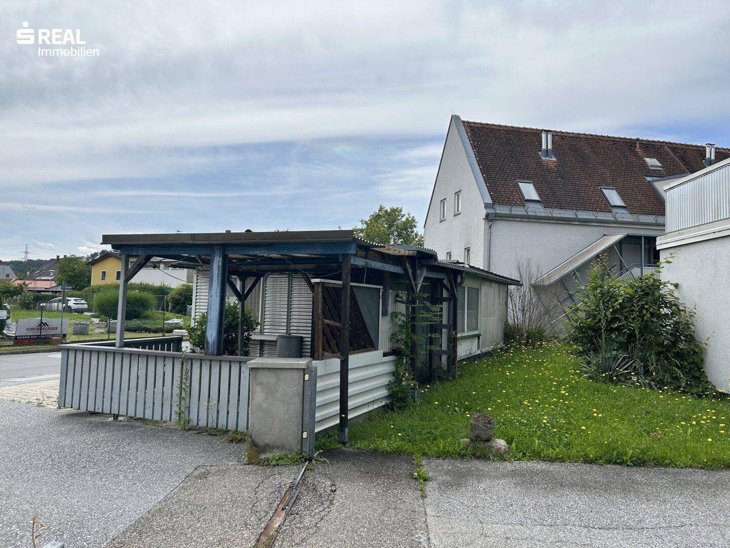 Ein kleines Gebäude mit einem überdachten Außenbereich und einem Wohnhaus im Hintergrund, umgeben von Gras.