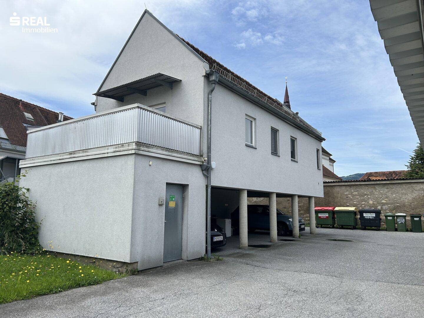 Seitenansicht eines Gebäudes mit einem überdachten Parkplatz im Erdgeschoss und einem Balkon darüber.