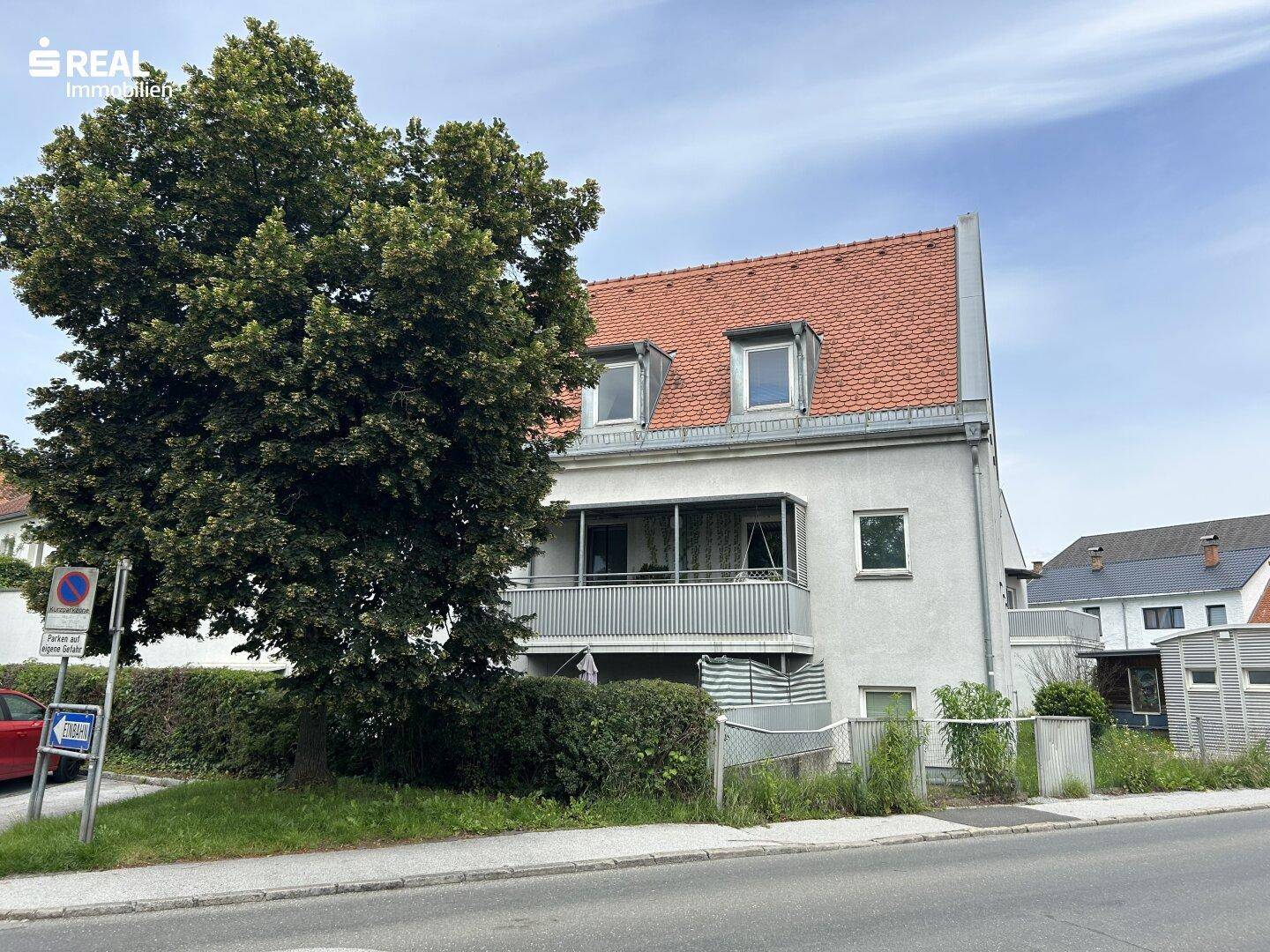 Vorderansicht eines Wohnhauses mit Satteldach, Balkon und einem großen Baum an der Straße.
