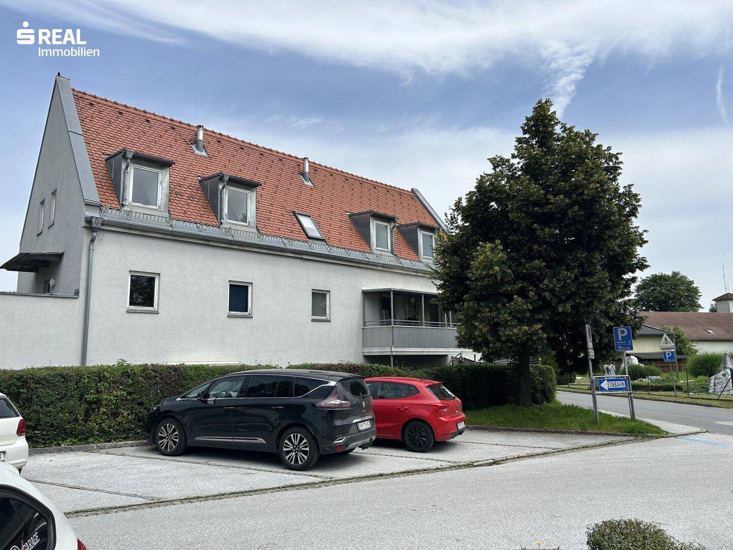 Wohngebäude mit Satteldach, Balkon und geparkten Autos auf einem Parkplatz an der Straße.