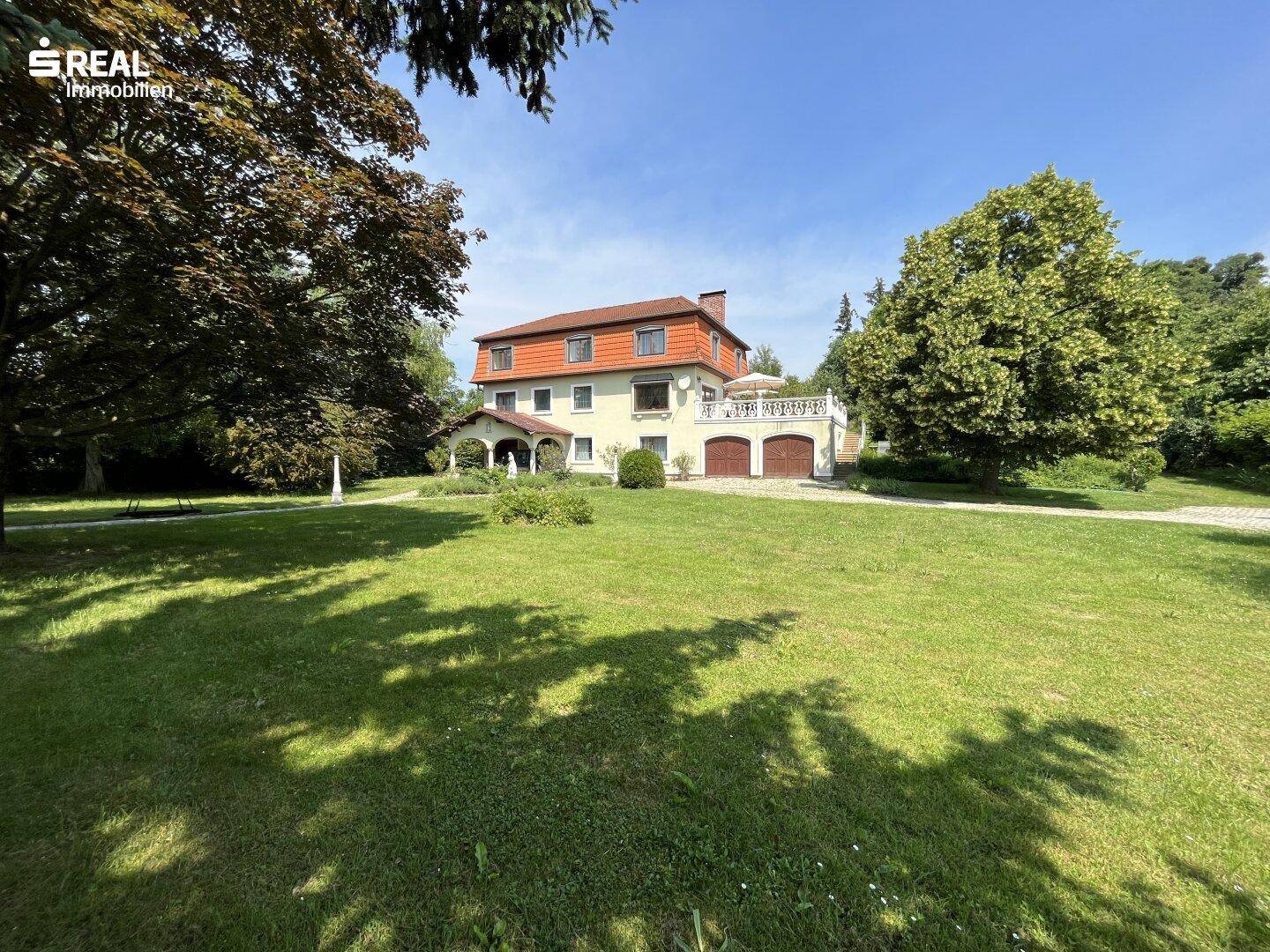 Weitläufiger Garten mit großem Haus und altem Baumbestand an einem sonnigen Tag.