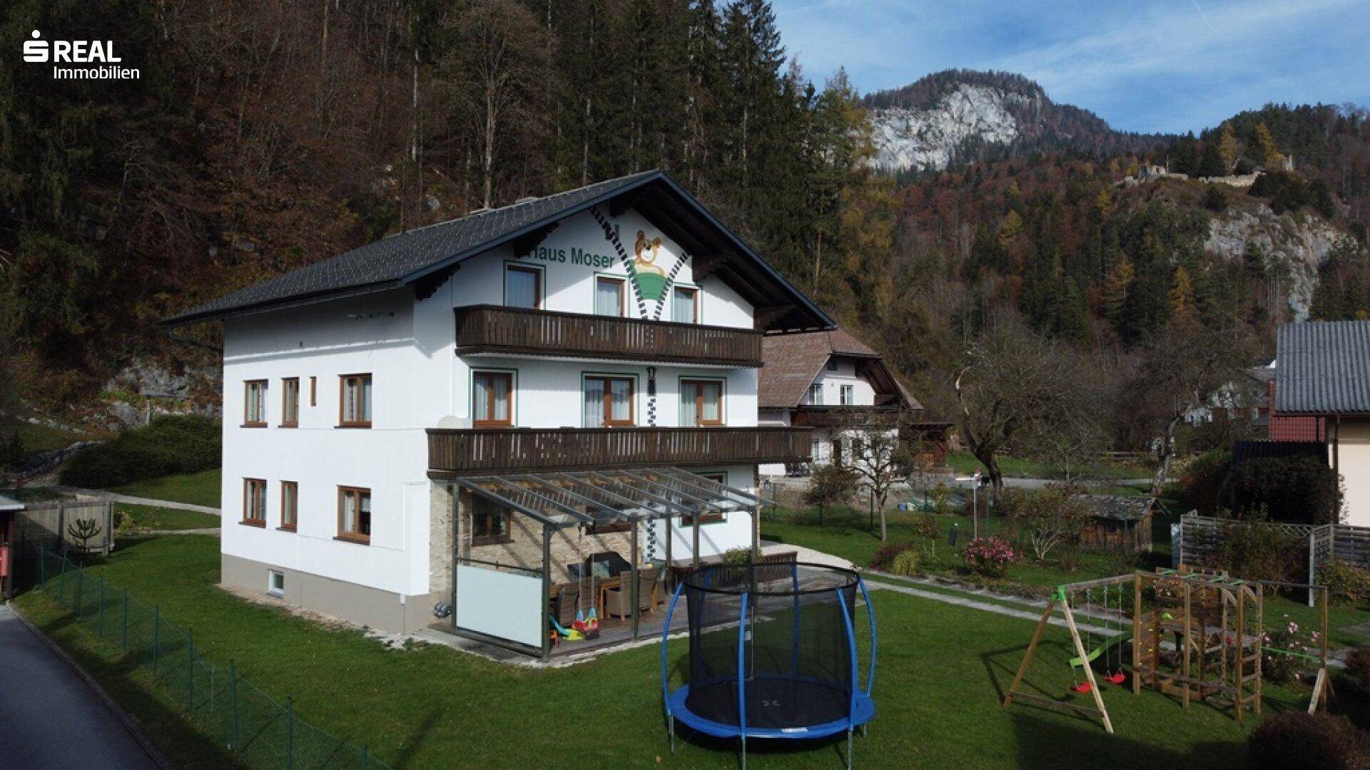 Traditionelles Haus Moser mit Balkonen und Spielplatz vor einer bewaldeten Bergkulisse.