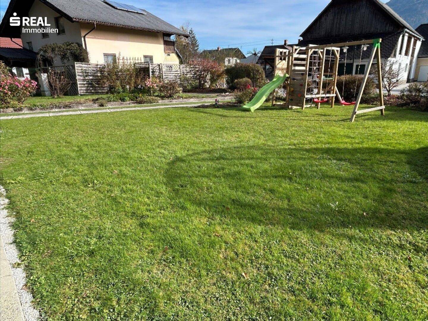 Großer Rasenbereich mit Kinderspielplatz, umgeben von weiteren Häusern und Hecken.