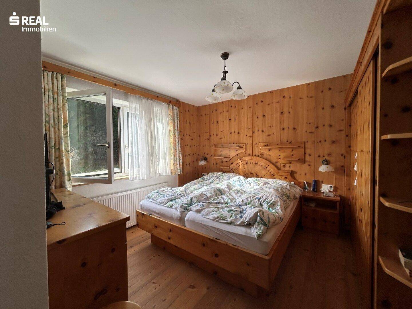 Gemütliches Schlafzimmer mit Holzwänden, Holzmöbeln und einem Fenster mit Blick ins Grüne.