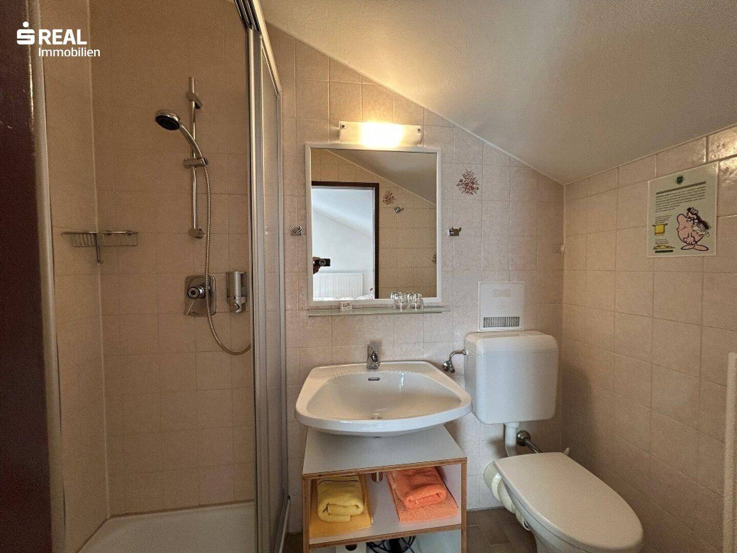 Gefliestes Badezimmer mit Dusche, Waschbecken und WC unter einer Dachschräge.