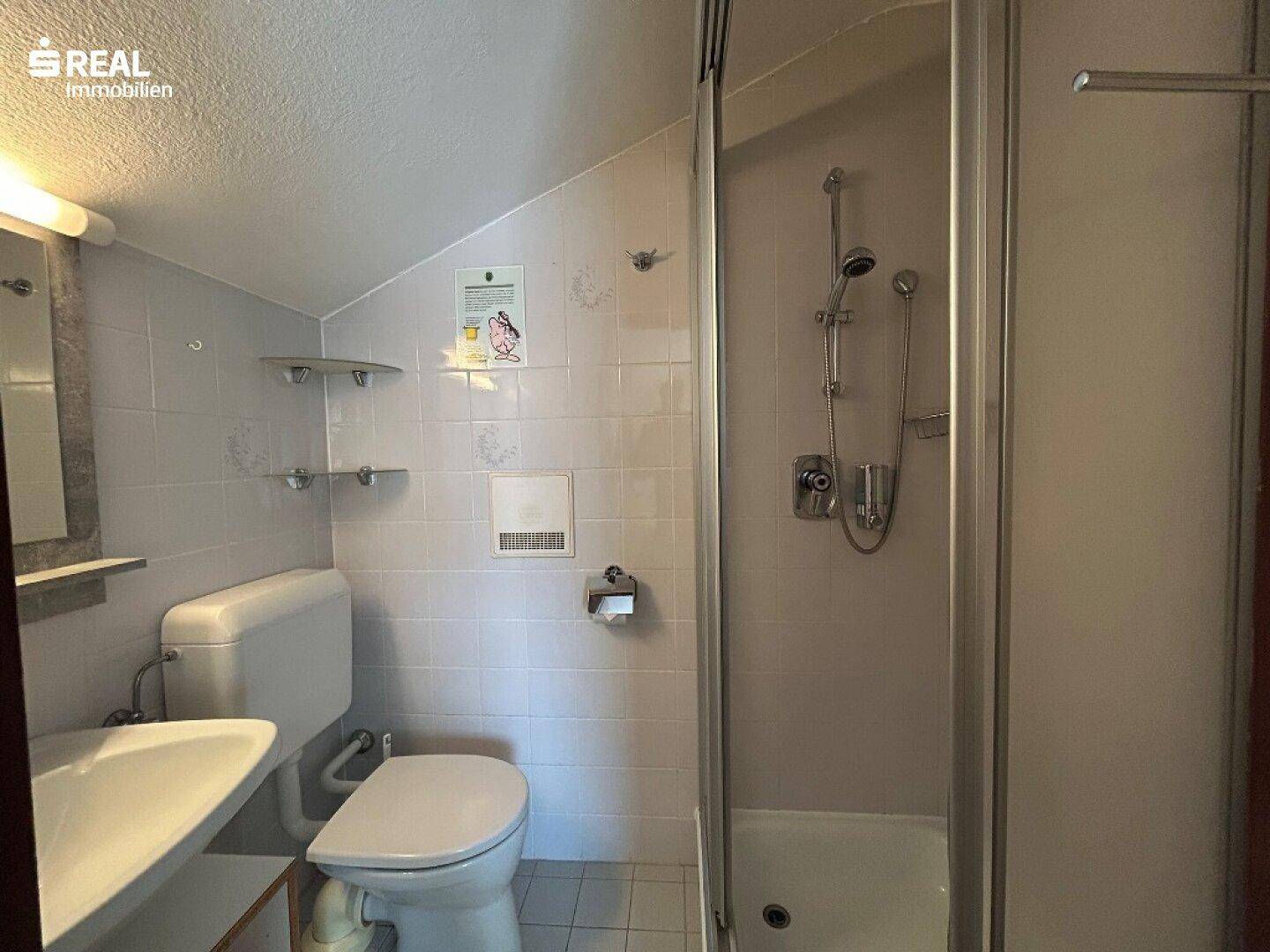 Kleines Badezimmer mit Dusche, WC und Waschbecken unter einer Dachschräge.