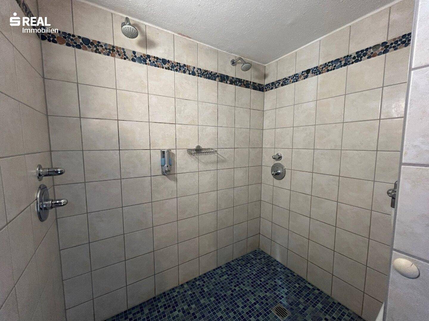 Geflieste Duschkabine mit zwei Duschköpfen und dekorativer Mosaikbordüre im Badezimmer.