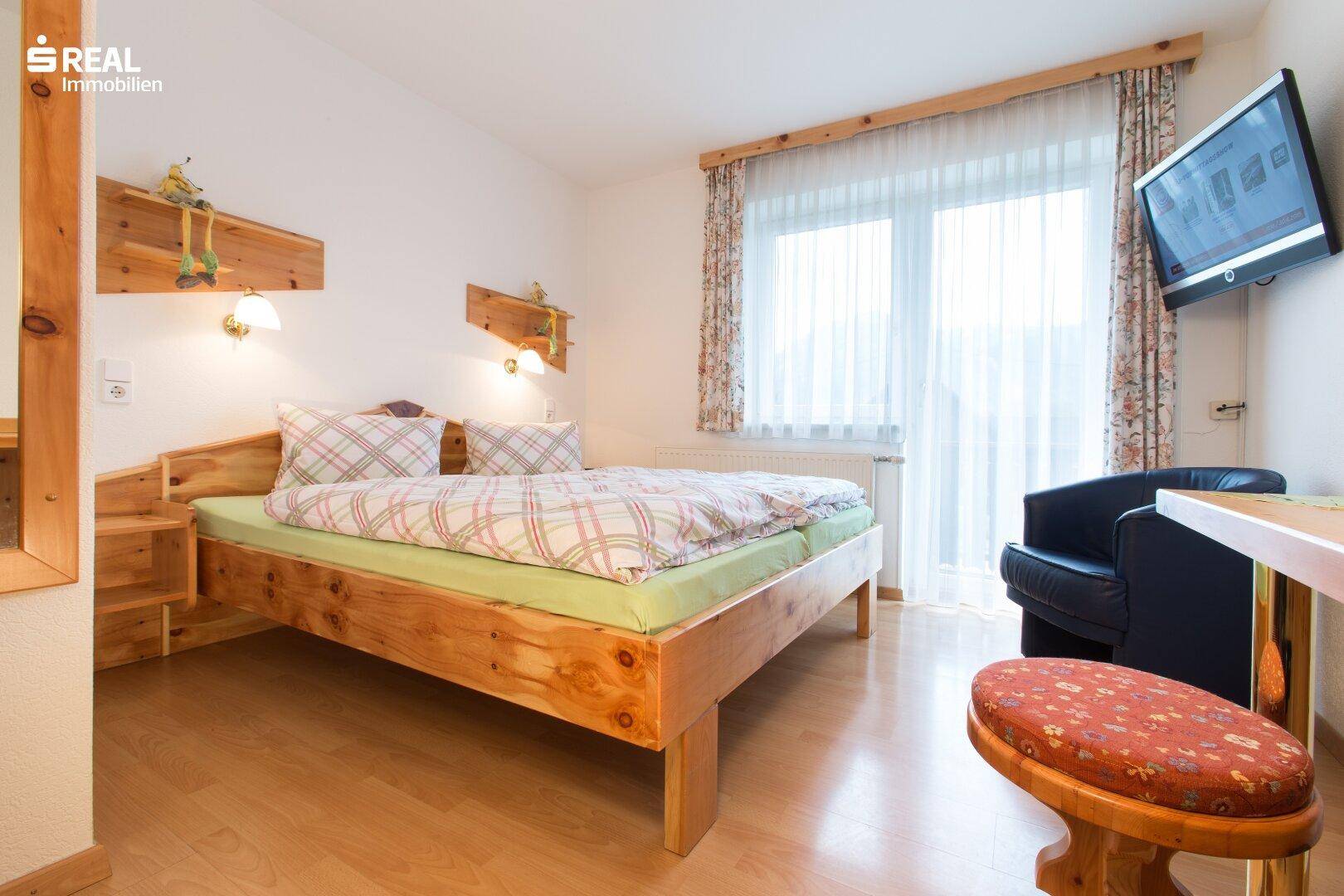 Schlafzimmer mit Holzfußboden, großem Fenster und einem an der Wand montierten Fernseher.