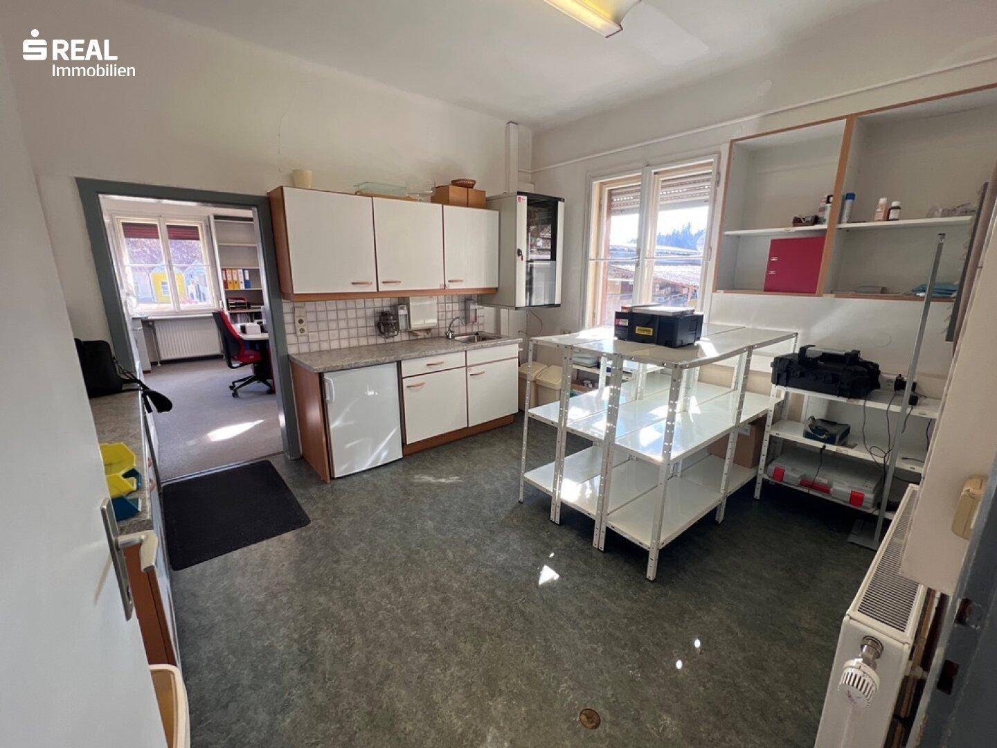 Einfache Teeküche mit Spüle, Kühlschrank und Regalen, angrenzend an ein Büro.