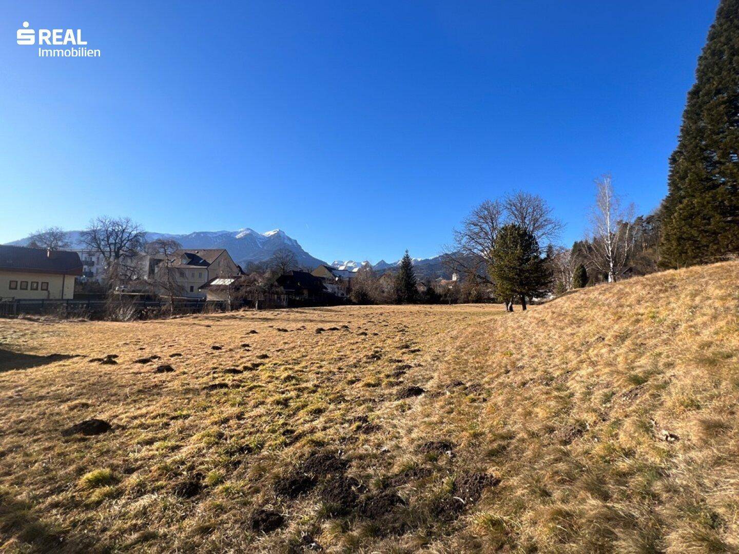 Sonniger Tag auf einem weitläufigen Grundstück mit Blick auf die Häuser und Berge im Hintergrund.