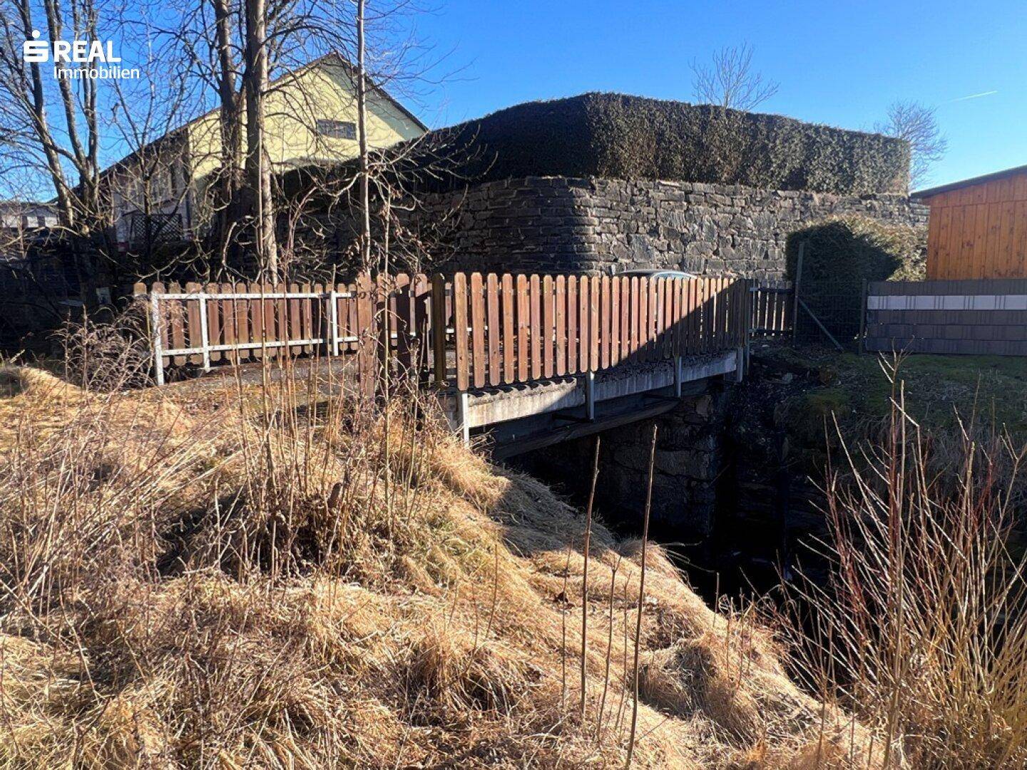 Brücke über einen Graben, die den Zugang zum Grundstück ermöglicht, umgeben von Natur.