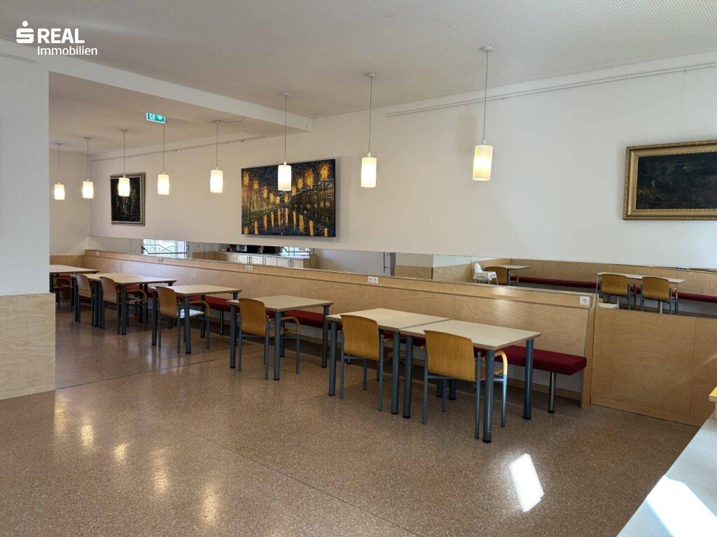 Langer Speisesaal mit Reihen von Tischen und Bänken, beleuchtet von Hängelampen.