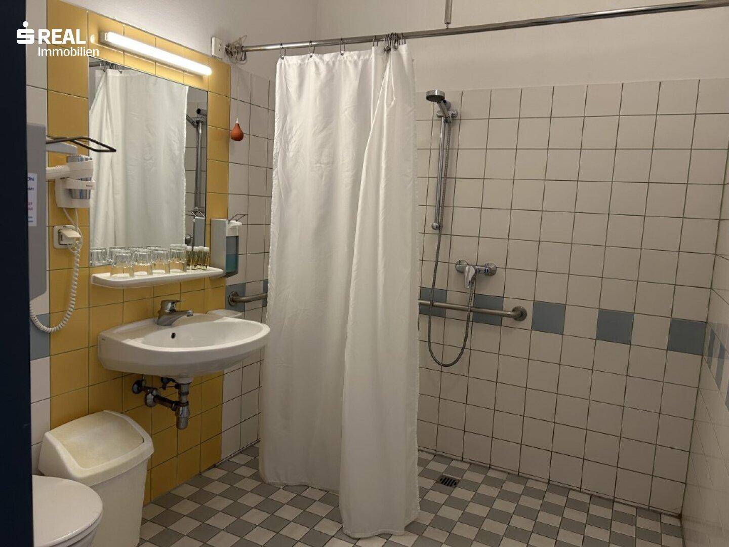 Badezimmer mit Dusche, Waschbecken und gelb-weißen Fliesen, ausgestattet mit Haltegriffen.