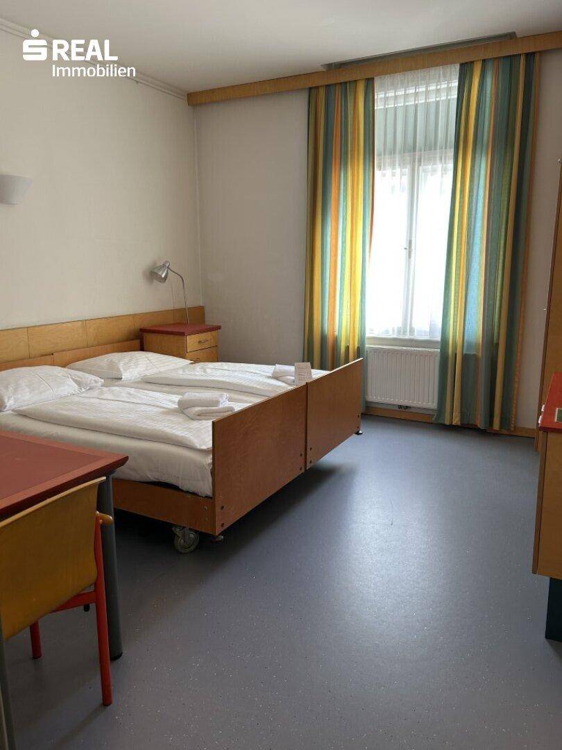 Schlafzimmer mit zwei Einzelbetten, Fenster mit bunten Vorhängen und Heizkörper.