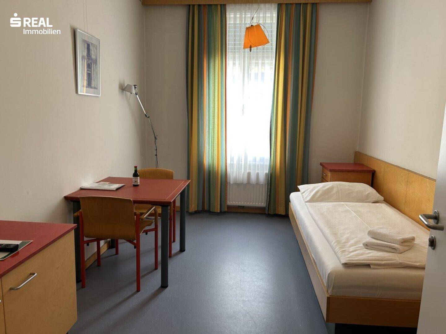 Einzelzimmer mit Bett, Schreibtisch und Stühlen, beleuchtet durch ein großes Fenster.