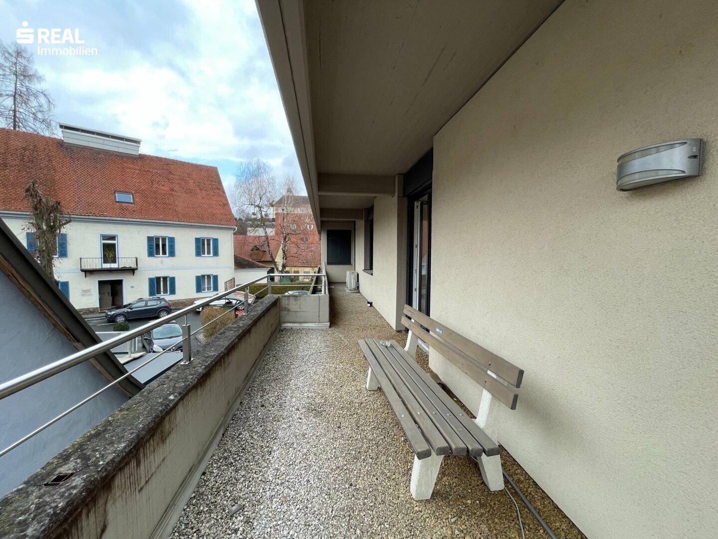 Langer Balkon mit Metallgeländer und einer Holzbank, bietet Blick auf die umliegenden Gebäude.