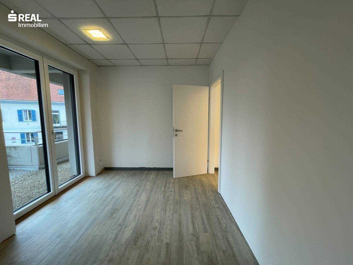 Leeres Büro mit großen Fenstern, die Zugang zu einem Außenbereich bieten, und hellem Laminatboden.