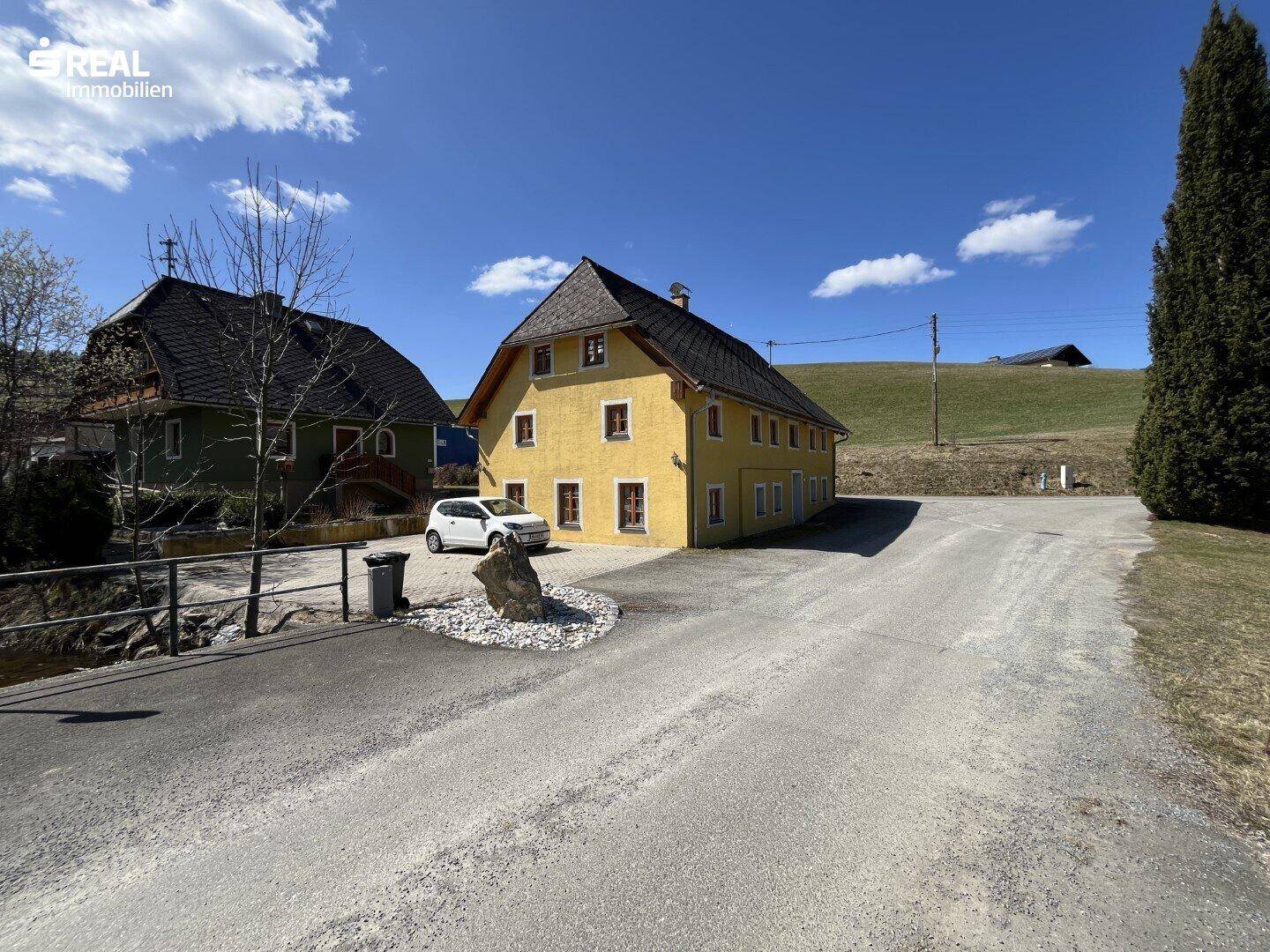 Gelbes Landhaus mit Schotterzufahrt und grünen Hügeln im Hintergrund unter blauem Himmel.