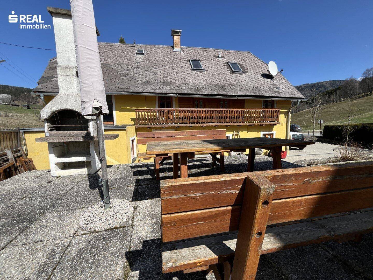 Große Terrasse mit Holztischen, Bänken und einem Grillbereich unter blauem Himmel.