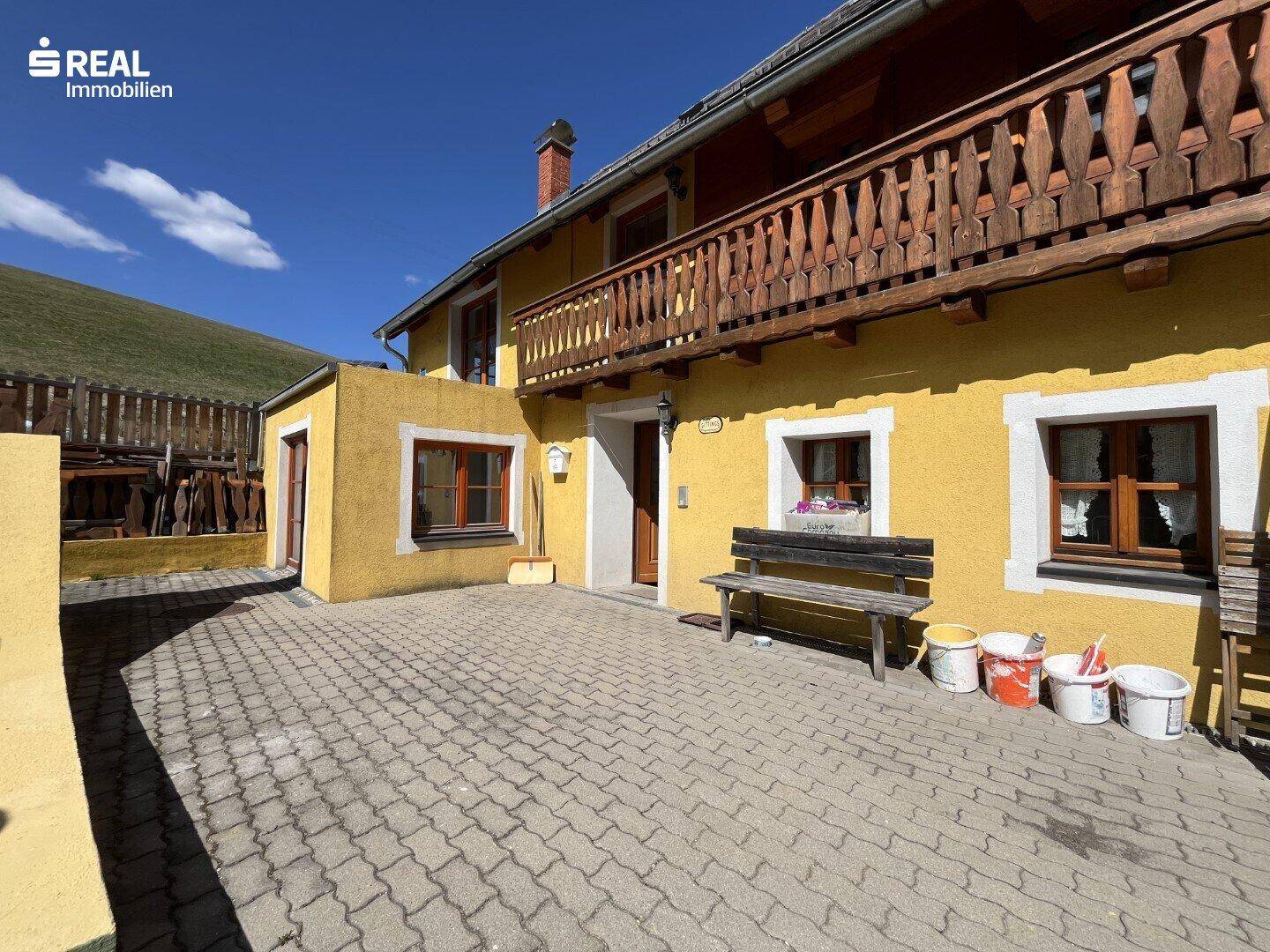 Gepflasterte Terrasse mit Holzbänken und Zugang zum Haus, umgeben von ländlicher Natur.