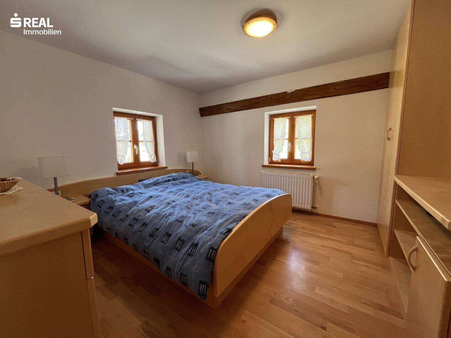 Helles Schlafzimmer mit Doppelbett, Holzmöbeln und zwei Fenstern für viel Tageslicht.