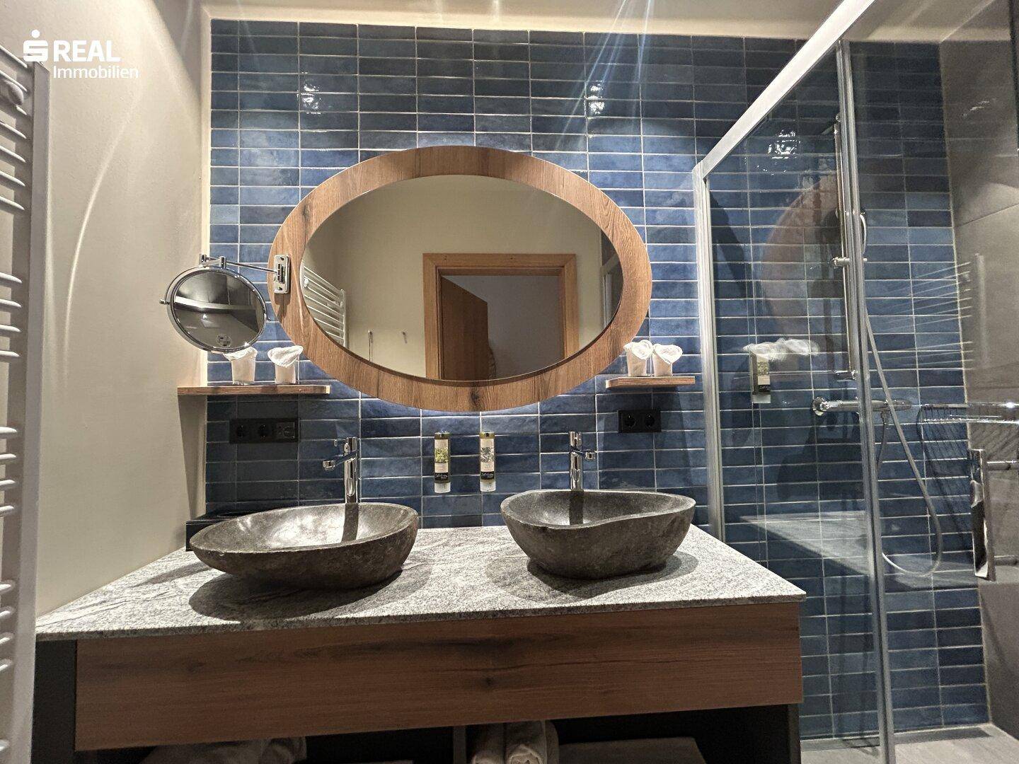 Modernes Badezimmer mit zwei Steinwaschbecken, ovalem Spiegel und blauen Fliesen an der Wand.