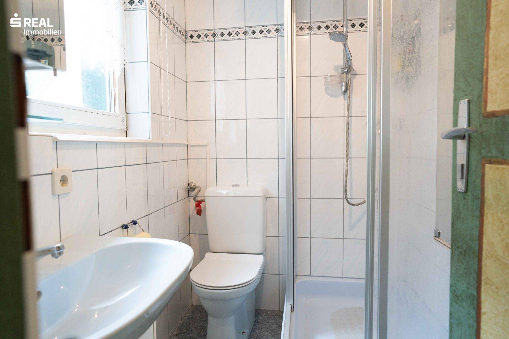 Helles Badezimmer mit Dusche, WC, Waschbecken und Fenster, ausgestattet mit weißen Fliesen.