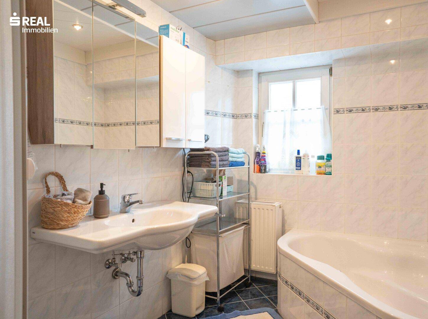 Funktionales Badezimmer mit Badewanne, Waschbecken, Spiegelschrank und Fenster.