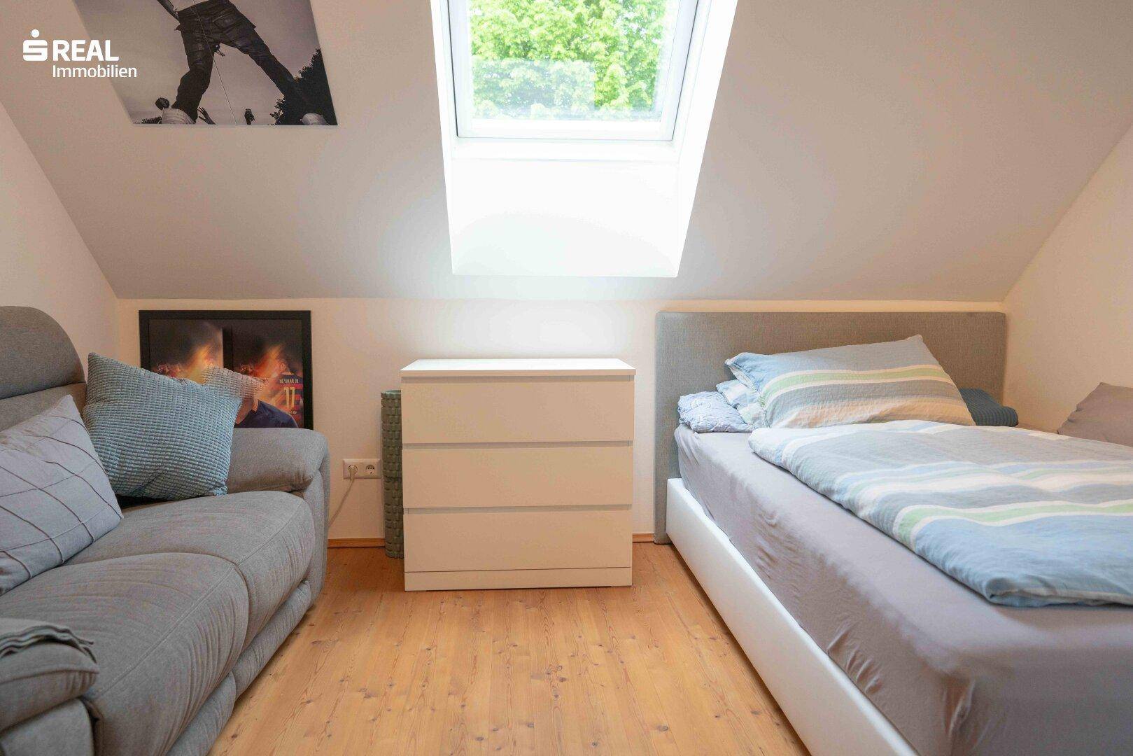 Helles Schlafzimmer im Dachgeschoss mit Einzelbett, Sofa und großem Dachfenster.