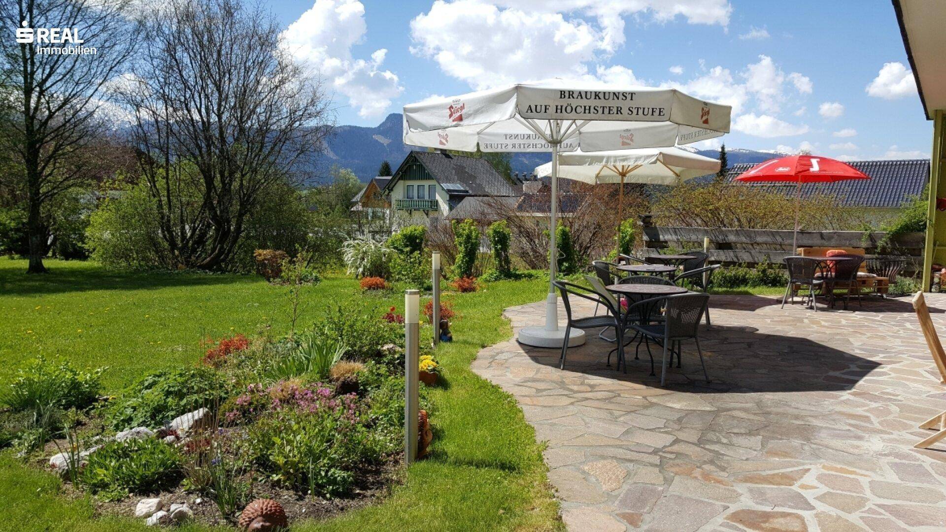 Gemütliche Außenterrasse mit Sitzgelegenheiten und Sonnenschirmen, umgeben von einem blühenden Garten und Bergblick.
