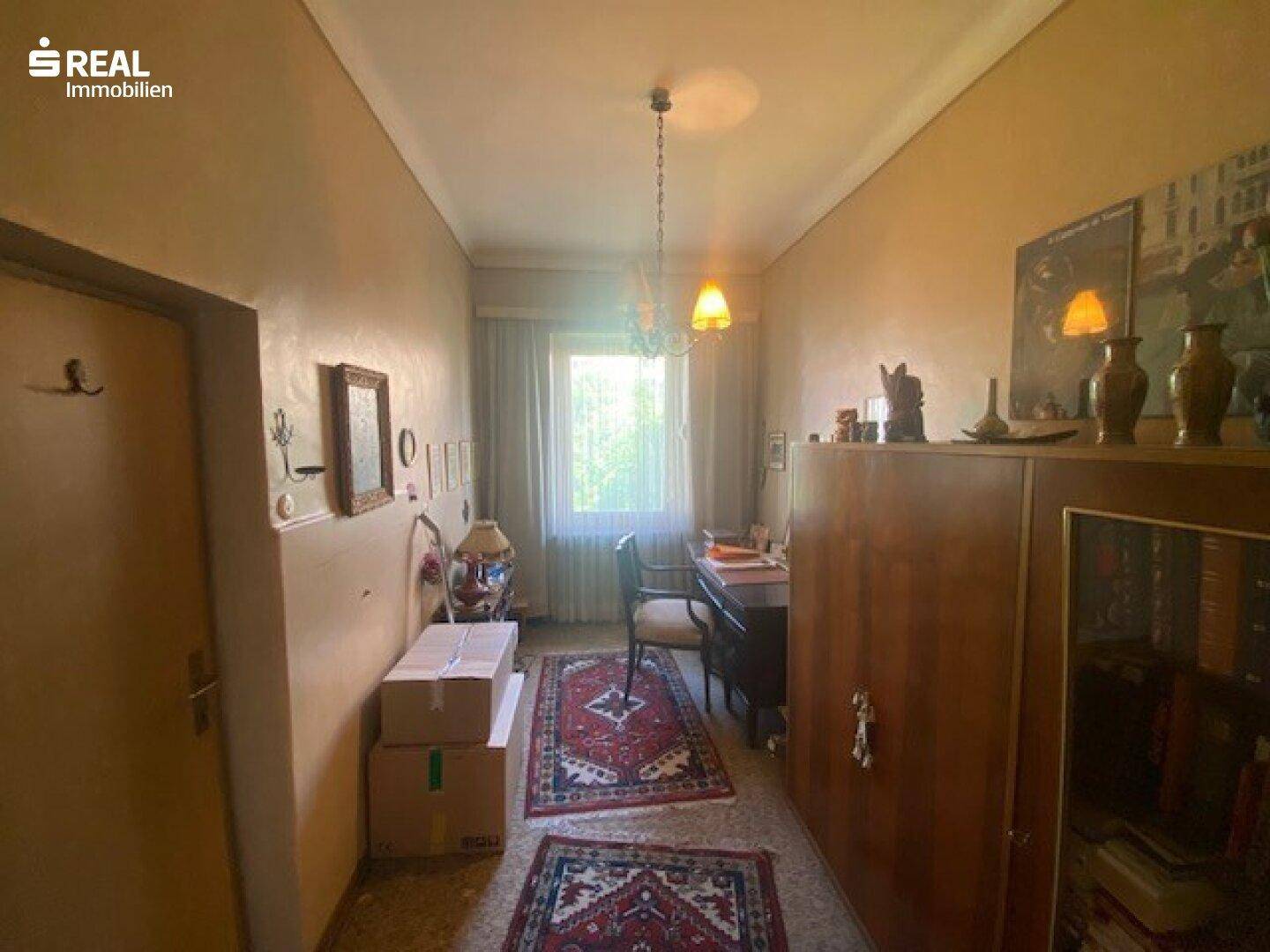 Schmaler Raum mit Fenster, Schreibtisch und Stauraum, möglicherweise ein Flur oder Büro.