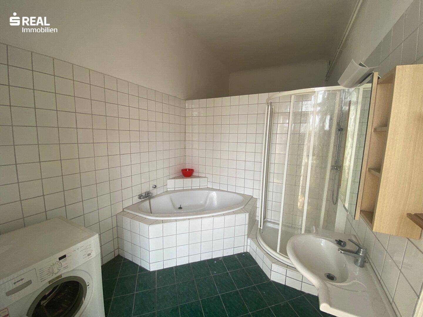 Badezimmer mit Eckbadewanne, Duschkabine und Waschmaschinenanschluss, gefliest in Weiß und Grün.