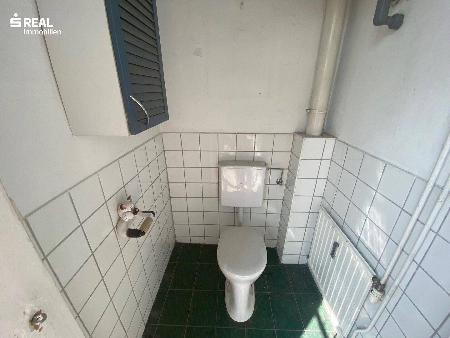 Kleines separates WC mit weißen Fliesen und grünem Boden, funktional und einfach gehalten.