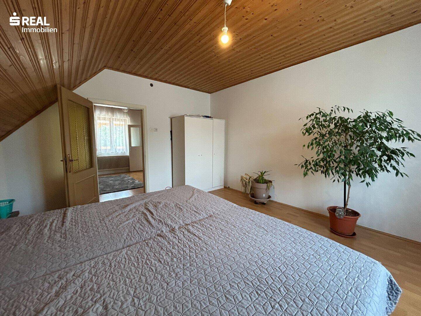 Schlafzimmer im Dachgeschoss mit Holzdecke, Blick auf den angrenzenden Raum und Pflanzen.