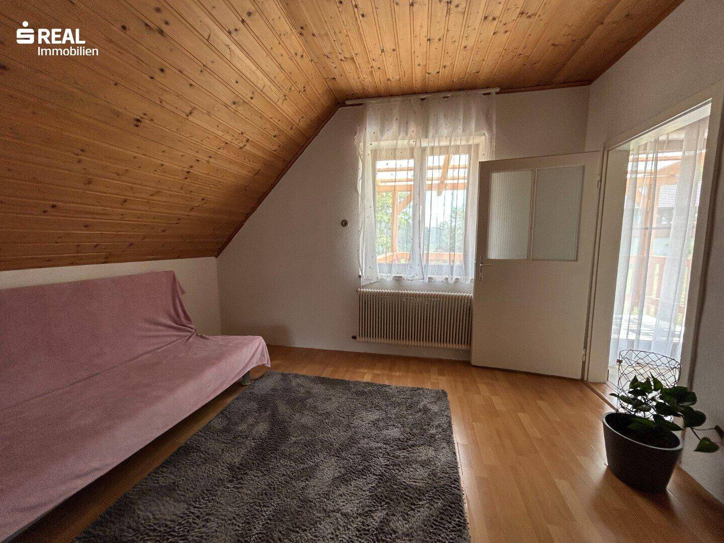 Helles Zimmer im Dachgeschoss mit Holzdecke, Fenster und Zugang zum Balkon.