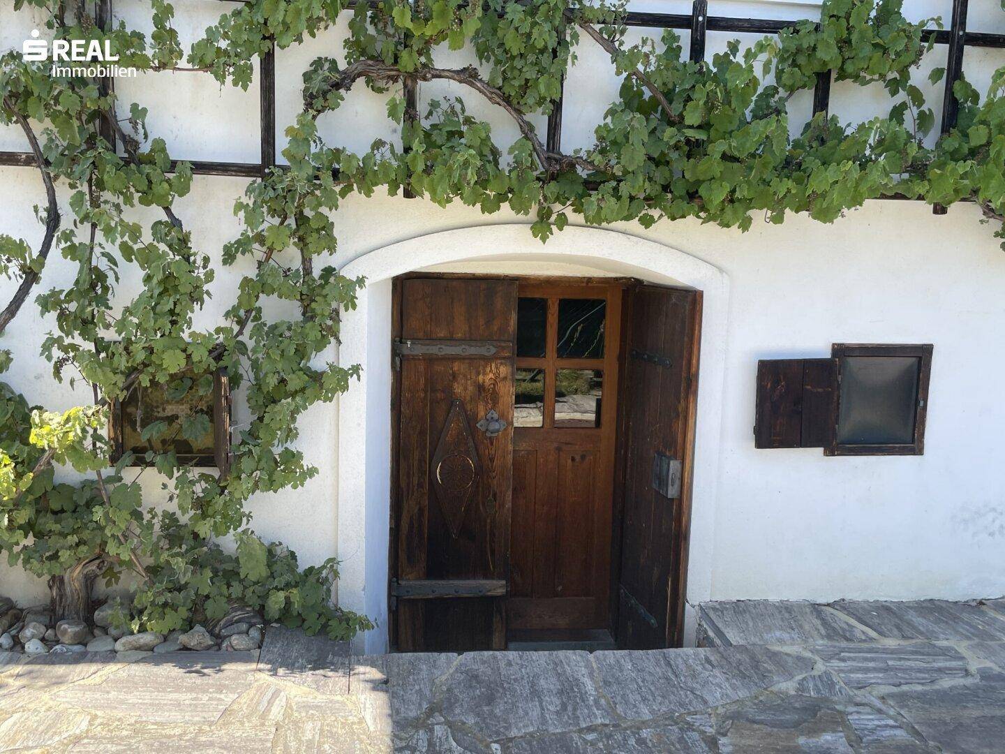 Rustikale Holztür als Eingang, umgeben von üppigen Weinreben und Steinplatten.