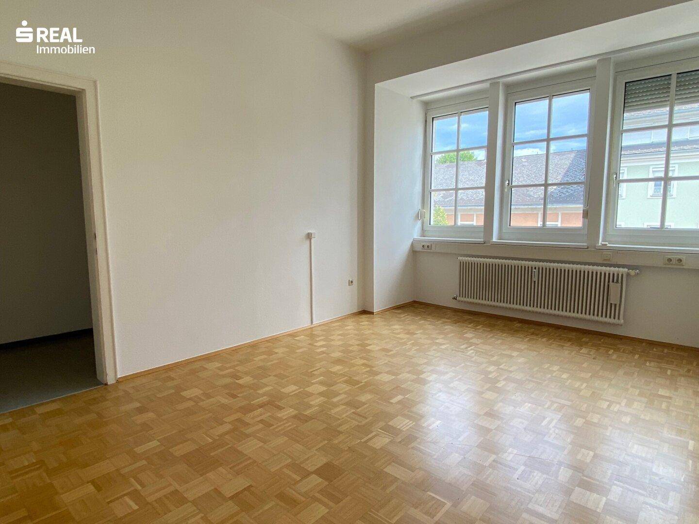 Heller Büroraum mit Holzparkett, großen Fenstern und Rollläden, die viel Tageslicht hereinlassen.