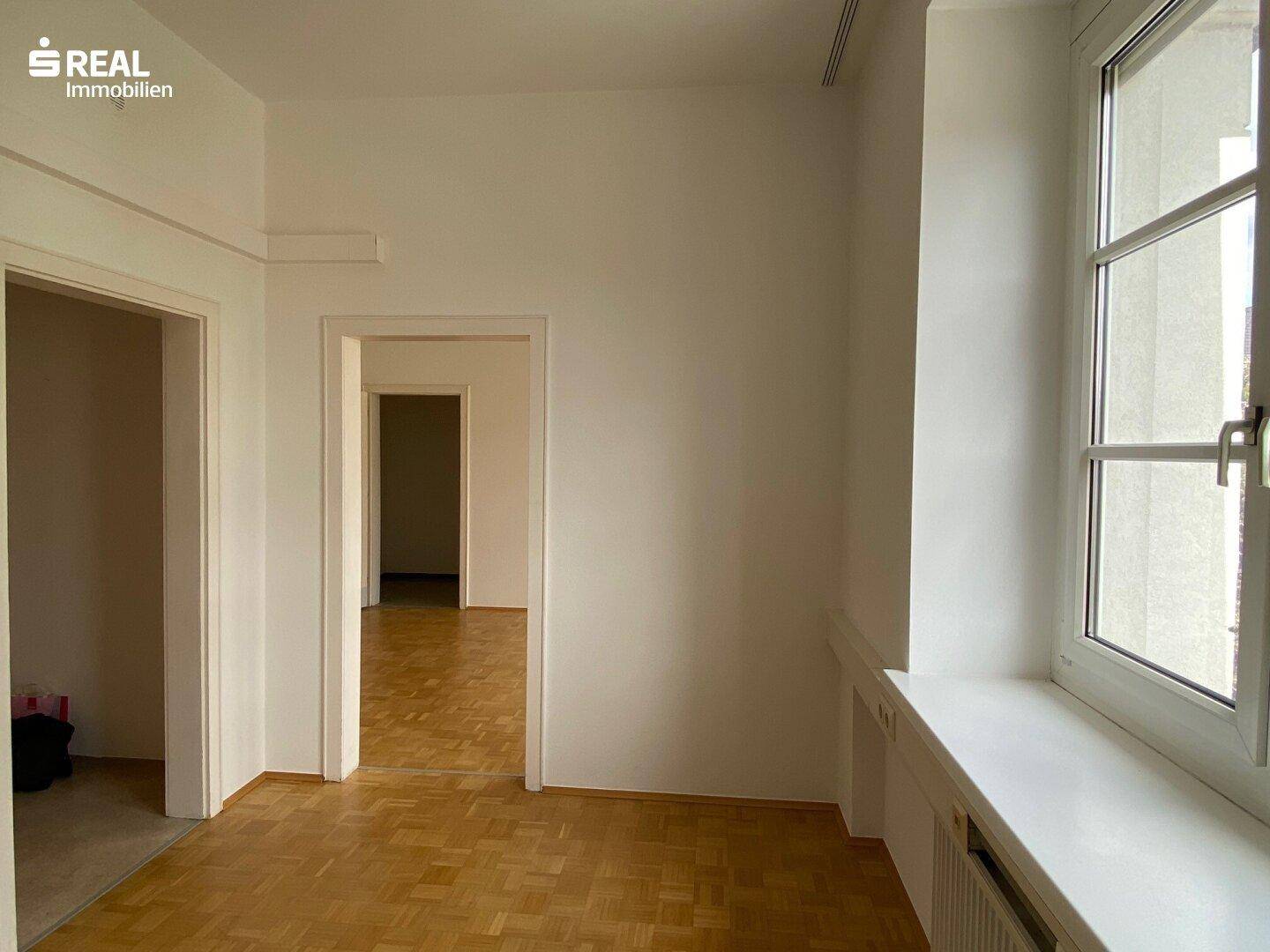 Flur oder Verbindungsbereich zwischen Büroräumen mit Parkettboden und natürlichem Lichteinfall durch ein Fenster.
